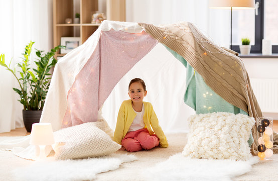 childhood and hygge concept - happy little girl sitting in kids tent at home