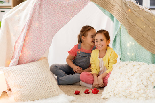 childhood and hygge concept - happy little girls playing tea party with toy crockery in kids tent at home and sharing secrets
