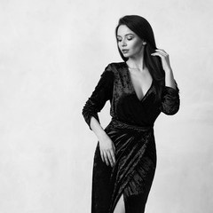 Fototapeta premium Beautiful young long haired brunette woman dressed in stylish long pale blue velvet dress with high split standing against white wall on background. Elegant female model wearing evening outfit.