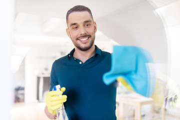 household and people concept - man in rubber gloves cleaning window with rag and spray cleaner at...