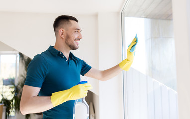 household and people concept - man in rubber gloves cleaning window with rag and spray cleaner at...
