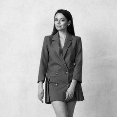 Pretty female model wearing gray mini blazer dress with four buttons and heeled shoes posing in studio. Beautiful woman in trendy outfit standing with crossed legs against white wall on background.