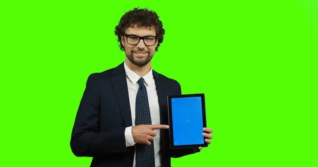 Caucasian good looking man in suit and tie posing with a black tablet computer in hands and pointing with his finger at it on the chroma key background. Green screen. Tracking motion