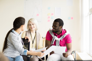 Fototapeta premium Two young women and black man talking about business smiling and sitting on chairs in light office