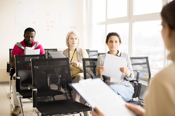 Two young women and black man holding papers in hands and sitting on chairs in light conference hall