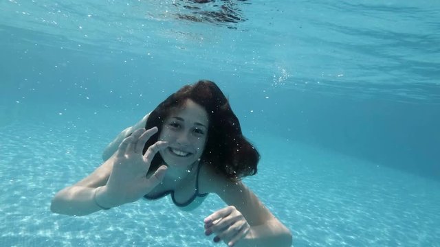 A teenage girl is swimming and playing under water in the pool, laughing and waving his arms. Blowing bubbles and looking at the camera. Action camera. 4K.