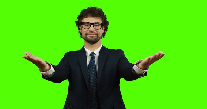 Young Caucasian businessman in glasses, suit and tie doing gesture like welcome with hands while standing on the green screen. Chroma key.