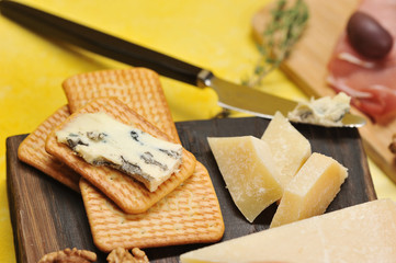 Cheese appetizer. Cheese with mold dorblu on the cracker and pieces of parmesan on a wooden board. In the frame a knife and slices of Parma ham. Close-up. Macro photography. Yellow background.