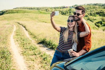 smiling stylish couple in sunglasses with coffee cup taking selfie on smartphone near car on rural meadow