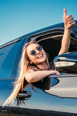smiling stylish woman in sunglasses leaning out from car window and doing thumb up gesture