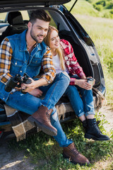 male tourist with binoculars and his smiling girlfriend sitting near with coffee cup on car trunk in field