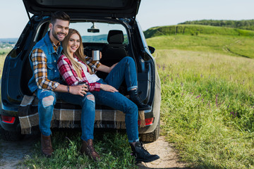 smiling couple with coffee cups sitting on car trunk in rural field