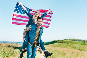 young man doing piggyback ride to girlfriend holding american flag on rural meadow, independence...
