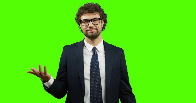 Good looking Caucasian businessman in glasses, suit and tie standing on the green screen and doing gesture so-so or fifty-fifty with a hand. Chroma key.