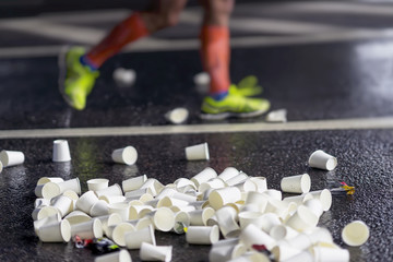 Pile of used disposable white cardboard cups for beverages and legs of runner on marathon at service point, selectiv focus, blur