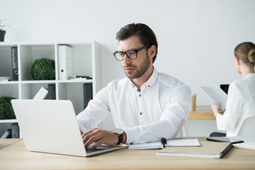 focused young businessman working with laptop at modern office with colleague sitting on background