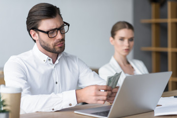 serious young businessman counting cash while his colleague looking at him blurred on background