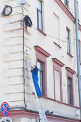 A young men in blue dress is staying on stair in outside. Worker is fixing a surveillance, security street camera to the wall of an old building.