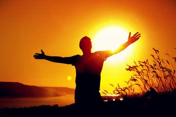Fototapeta premium Silhouette of relaxed man enjoying sunset time, raising hands to the sky. Travelling and vacation concept.