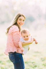 Fototapeta premium Young mother holding daughter in her arms. Beautiful Mother And Baby outdoors. Nature. Beauty Mum and her Child playing in Park together. Outdoor Portrait of happy family. Joy. Mom and Baby