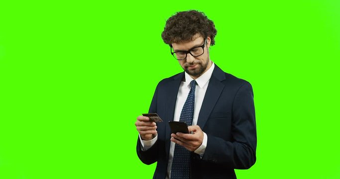 Attractive businessman in glasses and tie entering the credit card number on his smartphone while paying online and standing on the green screen background and working on the laptop. Chroma key.