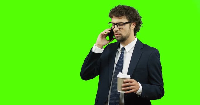 Attractive businessman in glasses and tie talking on his smartphone while drinking coffee to go on the green screen background. Chroma key.