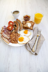 Flat lay of full English breakfast with fried eggs, bacon, sausages, beans and toasts. White wooden background.