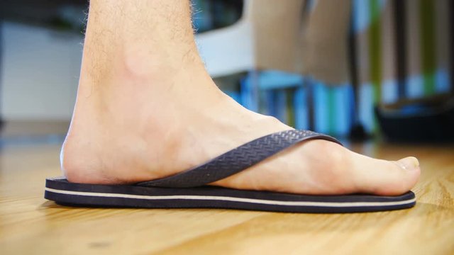Flat feet in flip-flops step in frame slow motion 4K. Low angle view of person's foot wearing flip-flops in focus while walking into frame stopping. Home dining room in the background out of focus.