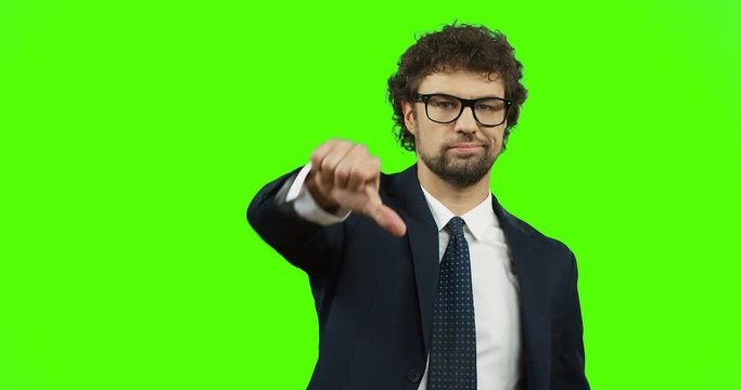 Attractive man in glasses, suit and tie giving his thumb down while standing on the green screen background. Chroma key.