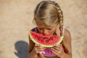 Portrait of a pretty blonde little girl eating watermelon on sandy beach on sunny day