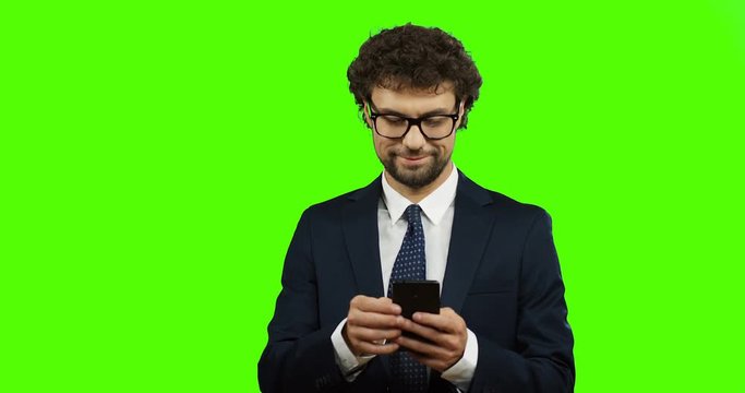 Attractive young man in the glasses, suit and tie using smartphone and then smiling to the camera. Green screen. Chroma key.