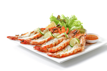 Grilled sliced black tiger prawns with baked potatoes, slice of lemons, fresh vegetables and chili sauce on white square plate, isolated on white background with shadow, low angle front side view.