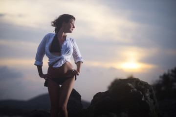 Sexy girl in a bikini stands on a rock against the sunset and looks into the distance