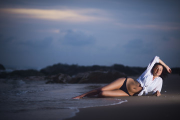 Sexy girl in bikini with a very beautiful body lying on the beach in the sea water at dusk against the sunset