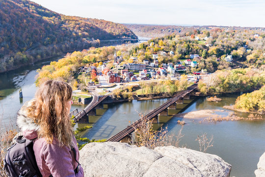 Hiker woman girl looking at cityscape overlook, colorful orange yellow foliage fall autumn forest with small village town by river in Harpers Ferry, West Virginia, WV