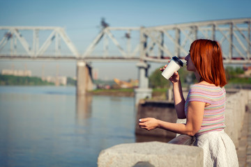 A young red-haired woman in a white romantic skirt, a pink top, drinks coffee and looks at the river on the city waterfront on a summer day
