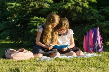 Two little girl friends schoolgirl learns sitting on a meadow in the park. Children with backpacks, books, notebooks.