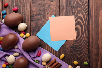 Chocolate Easter eggs with colorful candies and blank paper notes on wooden background
