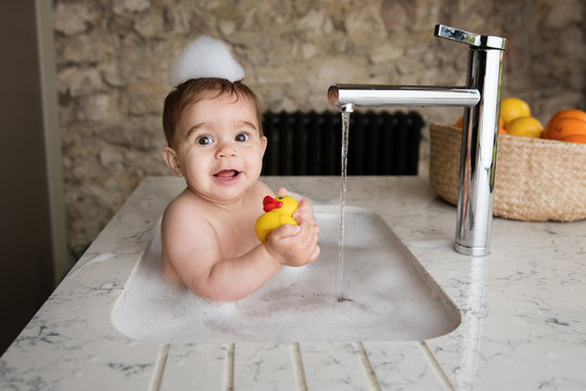 Baby taking bubble bath in kitchen sink holding rubber duck