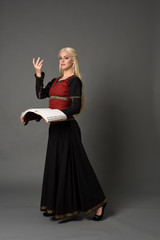  full length portrait of pretty blonde lady wearing  a red and black fantasy medieval gown, holding a book. standing pose on grey background.