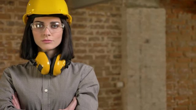 Young charming builder woman standing in earphones, glasses and helmet with crossed hands looking in camera, brick building background