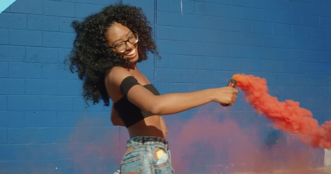 Beautiful young woman holding colorful smoke grenade dancing outside against blue wall 