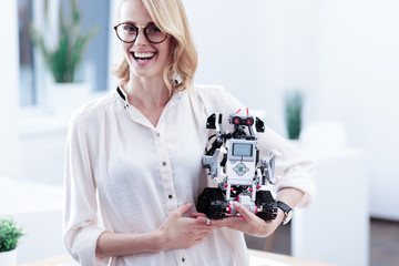Look here. Happy cheerful nice woman holding a robot and looking at you while being in a positive...