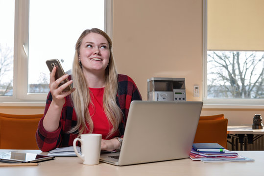 A smiling blonde woman sitting at a laptop and talking over a cell phone.