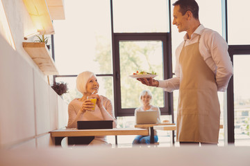 Obraz premium Here is your order. Pleasant handsome man serving a salad to his elderly customer while she sipping her juice and smiling at him