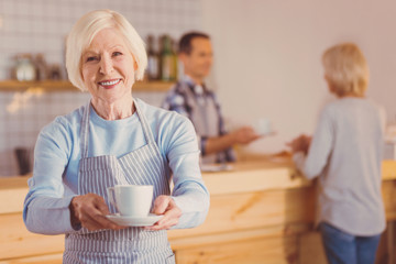 Enjoy your coffee. Beautiful elderly woman in an apron working as a barista in cafe and posing with a cup of coffee while smiling at the camera