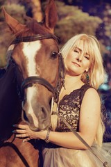 Portrait of a beautiful blonde woman in a chic dress with a horse walking in a pine forest in a stable