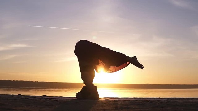 The girl does asana in yoga, slowly raises her legs in the rack on her head. Silhouette of a woman in the background of the shining sun