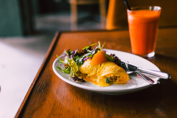 Morning breakfast, omelette with green salad on white plate with fresh orange carrot juice. Wooden table. Food photography, blogging concept.