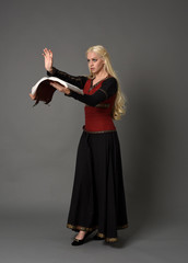  full length portrait of pretty blonde lady wearing  a red and black fantasy medieval gown, holding a book. standing pose on grey background.
